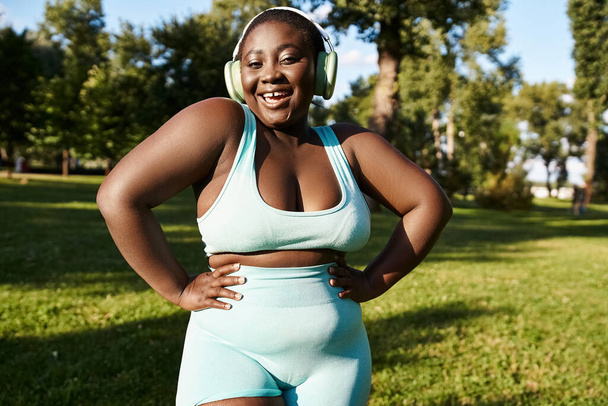 Афроамериканка в спортивной одежде стоит в траве, обнимаясь с положительным телом, слушая музыку через наушники. - Фото, изображение