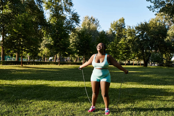 Una mujer afroamericana con curvas en sujetador deportivo verde y pantalones cortos azules sostiene una cuerda para saltar - Foto, Imagen