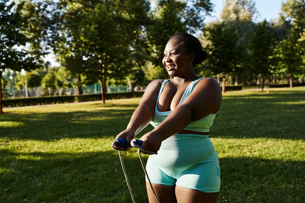Uma mulher afro-americana curvilínea em um sutiã esportivo verde e shorts azuis está se exercitando ao ar livre, segurando uma corda pulando. - Foto, Imagem
