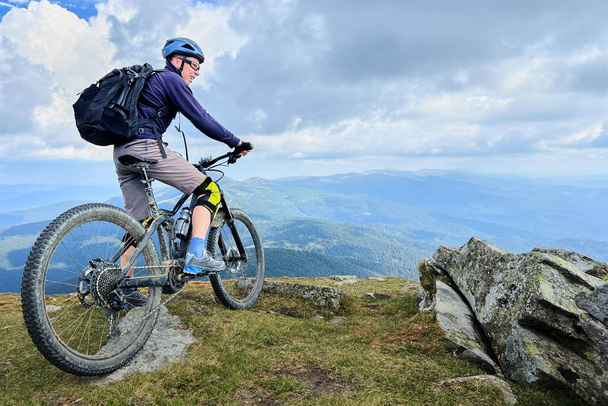 Велосипедист на улице на электрическом велосипеде. Мужской турист отдыхает на вершине холма, наслаждается красивым горным ландшафтом, носит шлем и рюкзак. Концепция активного отдыха. - Фото, изображение