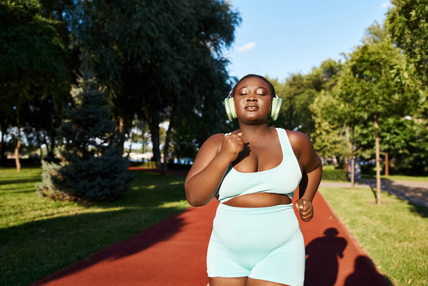 Μια καμπυλωτή Αφροαμερικανή γυναίκα με αθλητικά να τρέχει και να φοράει ακουστικά, απολαμβάνοντας την εξωτερική άσκηση ρουτίνας της. - Φωτογραφία, εικόνα