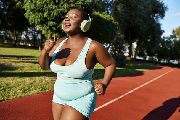 Una donna afroamericana in abbigliamento sportivo si sintonizza sulla musica con le cuffie mentre corre su una pista, incarnando la positività del corpo. - Foto, immagini