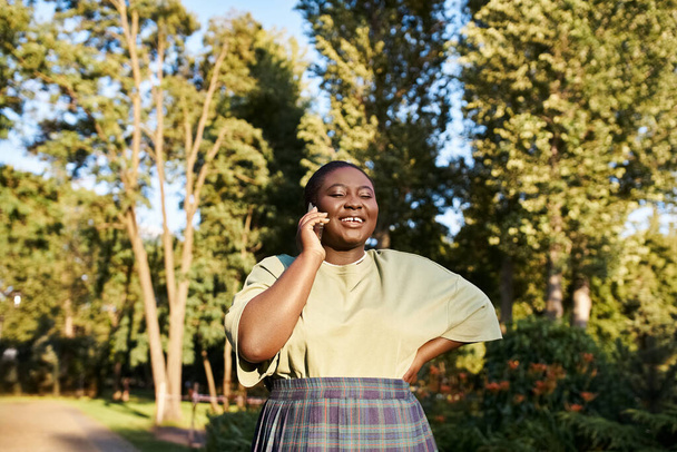 Plus-Size-Afroamerikanerin in lässiger Kleidung, umarmt Körperpositivität, plaudert an einem sonnigen Tag mit einem Handy in einem Park. - Foto, Bild