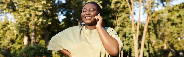 Eine Afroamerikanerin in lässiger Kleidung spricht auf einem Handy, während sie einen sonnigen Tag im Park genießt. - Foto, Bild
