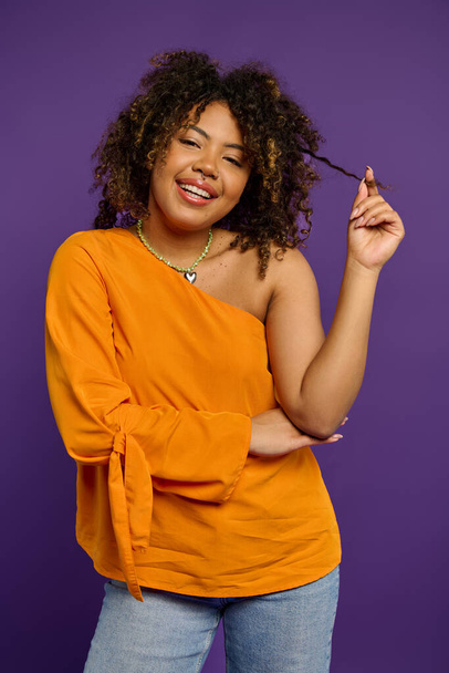 Κομψή Αφρο-Αμερικανίδα με πορτοκαλί μπλούζα που αγγίζει τα μαλλιά της. - Φωτογραφία, εικόνα
