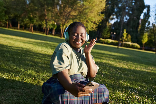 Uma mulher afro-americana plus-size desfrutando de música enquanto sentada na grama em traje casual, abraçando a positividade do corpo. - Foto, Imagem
