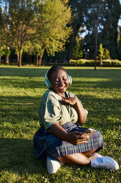 Büyük boy Afro-Amerikan bir kadın çimenlerde oturuyor, kulaklık takıyor, güneşli bir yaz gününde müzik dinliyor.. - Fotoğraf, Görsel