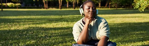 mujer en traje casual se sienta en la hierba, escuchando los auriculares, rodeado de naturaleza en un momento de verano tranquilo. - Foto, Imagen