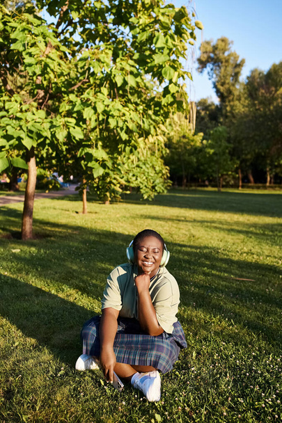 Ένα συν μέγεθος Αφροαμερικανή γυναίκα σε casual ρούχα κάθεται ειρηνικά στο γρασίδι σε μια ηλιόλουστη μέρα του καλοκαιριού. - Φωτογραφία, εικόνα