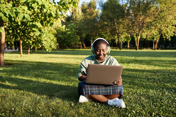 Een extra grote Afro-Amerikaanse vrouw in casual kleding zit in het gras met een laptop, genieten van een productieve werksessie buiten in de zomer. - Foto, afbeelding