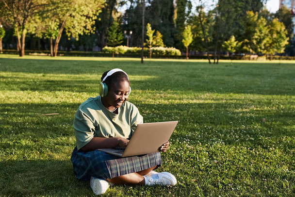 Плюс розмір афроамериканська жінка насолоджується природою під час роботи на ноутбуці в траві в сонячний день. - Фото, зображення