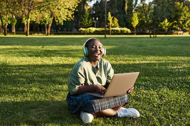 Büyük boy Afro-Amerikan bir kadın dizüstü bilgisayarıyla çimlerin üzerinde oturur ve yazın açık havanın tadını çıkarır.. - Fotoğraf, Görsel