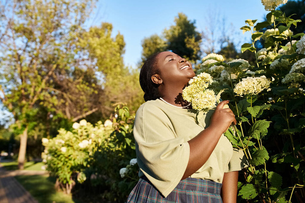 Una donna afroamericana plus size in abbigliamento casual si erge con grazia accanto a un cespuglio di fiori vibranti all'aperto in estate. - Foto, immagini