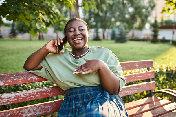 Συν μέγεθος Αφροαμερικανίδα γυναίκα σε casual ενδυμασία, κάθεται σε ένα παγκάκι έξω το καλοκαίρι, μιλώντας σε ένα κινητό τηλέφωνο. - Φωτογραφία, εικόνα