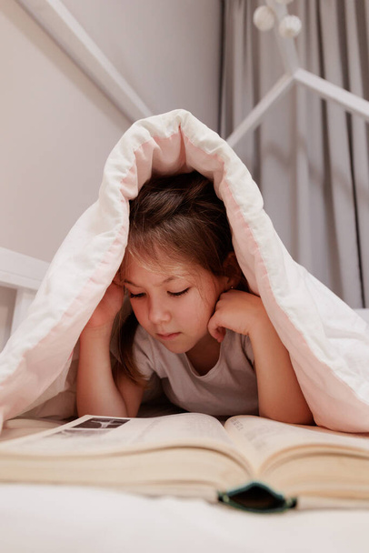 Küçük bir kızın portresi yatakta yatar ve battaniyenin altında kitap okur. - Fotoğraf, Görsel