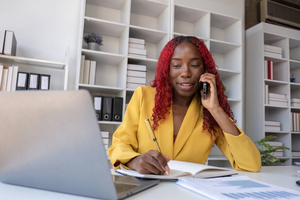 Mladá Afroameričanka mluví po telefonu a dělá si poznámky, když pracuje v kanceláři. - Fotografie, Obrázek