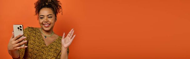 Μια όμορφη Αφρο-Αμερικανίδα με μοντέρνα ενδυμασία που κρατάει ένα κινητό μπροστά από έναν πορτοκαλί τοίχο.. - Φωτογραφία, εικόνα