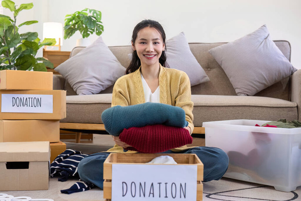 słodkie Azji kobieta siedzi obok kanapa w salonie sortowanie niechciane ubrania dla darowizny.. - Zdjęcie, obraz