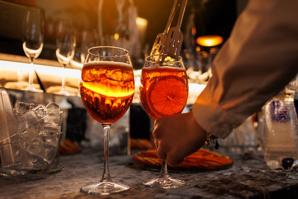 Barman maakt twee glazen cocktail Aperol spritz op bar teller, voegt verse sinaasappelschijfjes toe. Typische Italiaanse alcoholische drank, aperitief gemaakt van Prosecco mousserende witte wijn en ijsblokjes - Foto, afbeelding