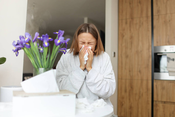 Mujer joven enferma que sufre de alergia, fiebre o gripe sopla la nariz y estornudos en los tejidos de papel, se sienta a la mesa en la cocina casera. Chica con secreción nasal, alergia al polen, gripe, resfriado, rinitis, sinusitis - Foto, Imagen