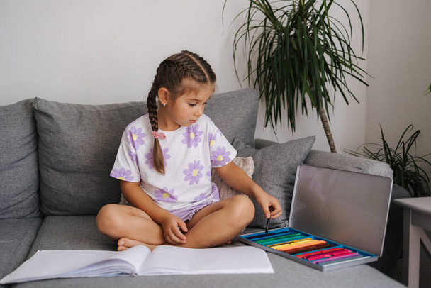 Cute little girl siedzi na kanapie i malowanie przez kolorowe ołówki. Nastrój domu. Wysokiej jakości zdjęcie - Zdjęcie, obraz