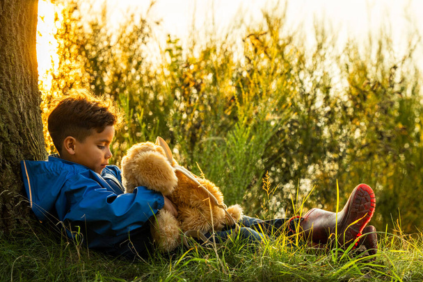 Obraz krásně zachycuje mladého chlapce v zamyšleném okamžiku, jak sedí se svým plyšovým medvídkem u stromu a dívá se na jezero zalité teplým světlem zapadajícího slunce. Kluci. - Fotografie, Obrázek