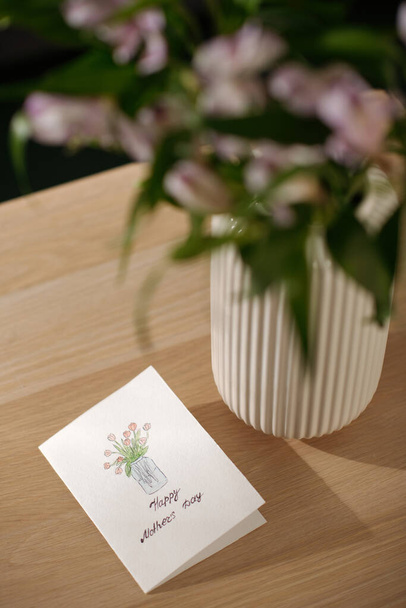 Część drewnianego stołu z ręcznie robioną pocztówką z pozdrowieniami na Dzień Matki i rysunkiem kilku czerwonych tulipanów w wazonie - Zdjęcie, obraz