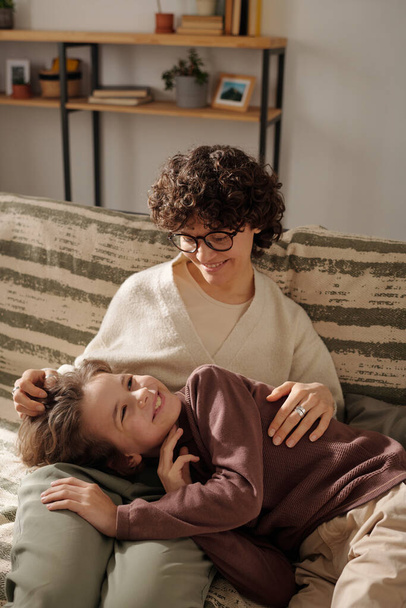 Heureuse jeune femme brune aux lunettes jouissant de jouer avec son adorable fils tout en se relaxant sur le canapé le week-end - Photo, image