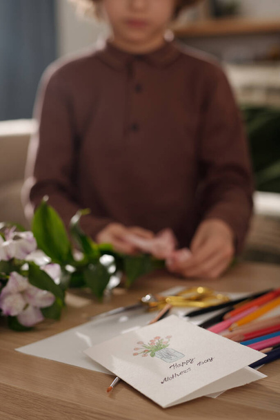 Tarjeta de felicitación hecha a mano con flores dibujadas en jarrón acostado en la mesa con lápices de colores contra el niño atando ramo para madre con cinta - Foto, imagen