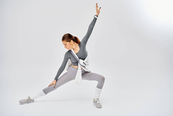 Eine sportliche junge Frau in grau-weißer Kleidung übt anmutig eine Yoga-Pose auf grauem Hintergrund. - Foto, Bild