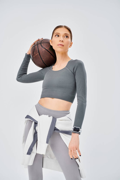 Eine sportliche junge Frau hält selbstbewusst einen Basketball in der rechten Hand und zeigt auf grauem Hintergrund ihr sportliches Können. - Foto, Bild
