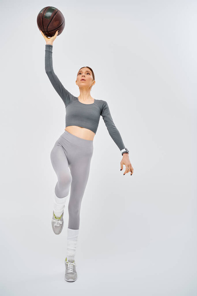 Aktif giyinmiş genç bir kadın gri bir arka planda kendine güvenen bir basketbol topunu havada tutuyor.. - Fotoğraf, Görsel