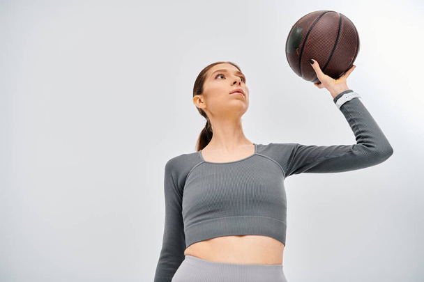 Eine sportliche junge Frau in aktiver Kleidung hält vor grauem Hintergrund einen Basketball in die Höhe. - Foto, Bild