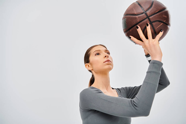 Sportowa młoda kobieta w aktywnym ubraniu trzymająca koszykówkę w powietrzu na szarym tle. - Zdjęcie, obraz