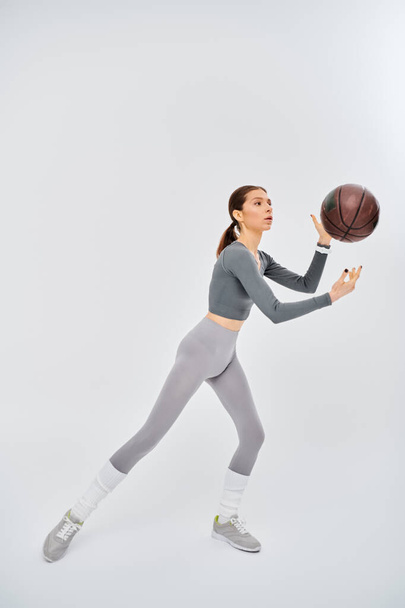 Een sportieve jonge vrouw houdt vol vertrouwen een basketbal in haar rechterhand, gekleed in actieve kleding tegen een grijze achtergrond. - Foto, afbeelding