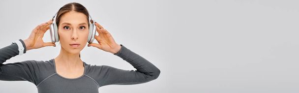 Αθλητική νεαρή γυναίκα σε ενεργό φορούν απολαμβάνοντας τη μουσική μέσω ακουστικών, τα χέρια ψηλά σε μια χαλαρή στάση σε ένα γκρι φόντο. - Φωτογραφία, εικόνα