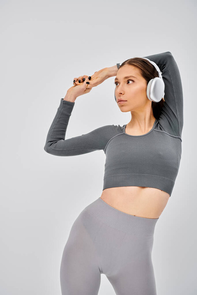 Una joven deportista en ropa activa hace una pose mientras escucha música a través de auriculares sobre un fondo gris. - Foto, imagen