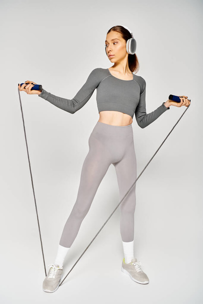 Mujer joven deportiva en ropa activa, sosteniendo saltar la cuerda, escuchando música sobre fondo gris. - Foto, Imagen