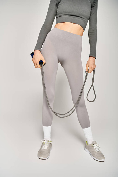Una joven deportista en ropa activa sosteniendo una cuerda de salto en sus manos sobre un fondo gris. - Foto, Imagen