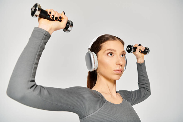Una joven deportista con auriculares, sosteniendo dos mancuernas, exudando fuerza y determinación sobre un fondo gris. - Foto, imagen