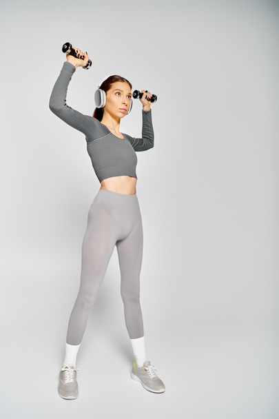 A sporty young woman in active wear is exercising with dumbbells on a grey background. - Photo, Image