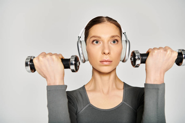 Eine sportliche junge Frau in aktiver Kleidung trägt Kopfhörer und hält zwei Kurzhanteln. - Foto, Bild