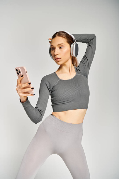 Sportovní mladá žena v aktivním oblečení, drží mobilní telefon a nosí sluchátka, zapojena do telefonátu na šedém pozadí. - Fotografie, Obrázek