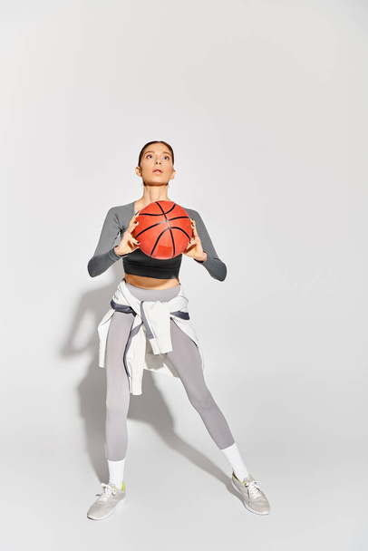 Egy sportos fiatal nő elegánsan jobb kezében tartja a kosárlabdát, szürke háttér mellett.. - Fotó, kép