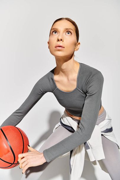 Sportovní mladá žena v aktivním oblečení drží basketbal v pravé ruce na šedém pozadí. - Fotografie, Obrázek