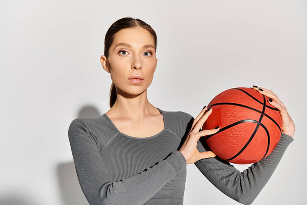 Een sportieve jonge vrouw in actieve kleding houdt sierlijk een basketbal in haar handen op een grijze achtergrond. - Foto, afbeelding
