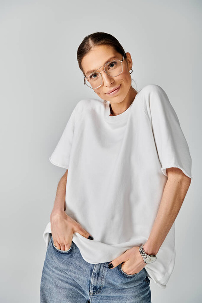 Μια νεαρή γυναίκα με λευκό μπλουζάκι και γυαλιά ποζάρει σε γκρι φόντο, αποπνέοντας κομψότητα και αυτοπεποίθηση. - Φωτογραφία, εικόνα
