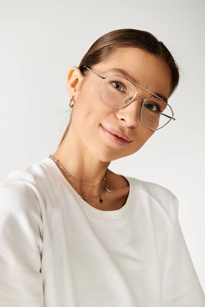 Μια νεαρή γυναίκα φορώντας γυαλιά και ένα λευκό πουκάμισο αποπνέει μια αίσθηση ηρεμίας σε ένα γκρίζο φόντο. - Φωτογραφία, εικόνα