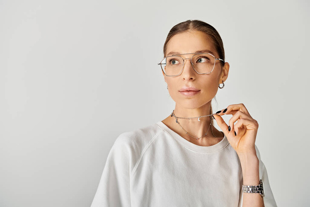 Una joven con gafas y una camisa blanca posa sobre un fondo gris, exudando una sensación de serena confianza. - Foto, Imagen