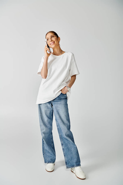 Une jeune femme vêtue d'une chemise blanche et d'un jean parlant sur un téléphone portable sur fond gris. - Photo, image